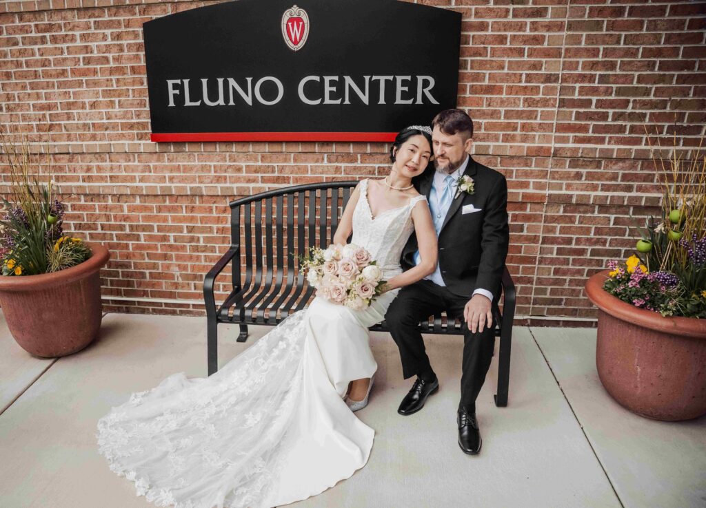 A bride and groom sit on a metal bench near the Fluno Center