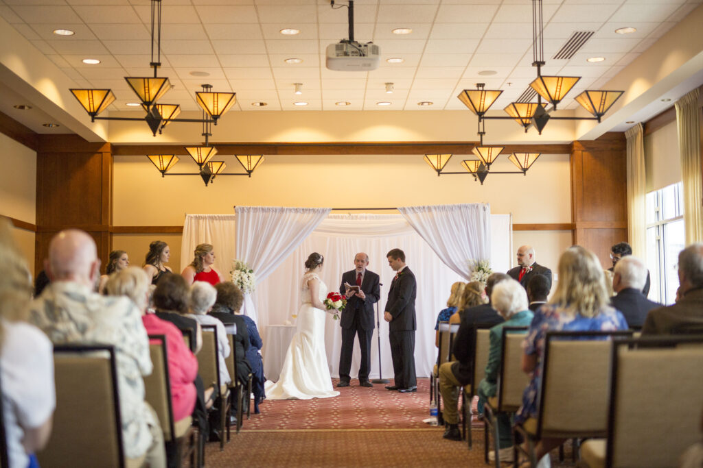 Wedding Ceremony seen from the aisle: seated guests watch a bride, groom, and officiant
